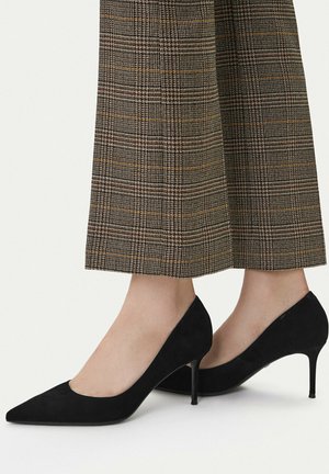 Décolleté in camoscio nero con punta a punta indossate con pantaloni a gamba larga a quadri marroni, mettendo in evidenza le calzature e la parte inferiore della gamba.