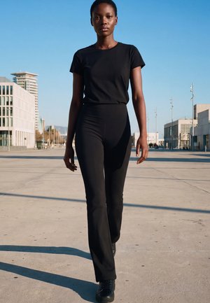 Tânără în tricou și pantaloni negri mergând cu încredere pe o piață urbană cu clădiri moderne și cer albastru limpede.