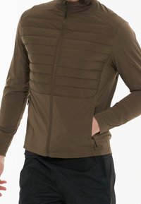 Man wearing an olive green quilted jacket with a front zipper, side pockets, and black pants, hand in jacket pocket.