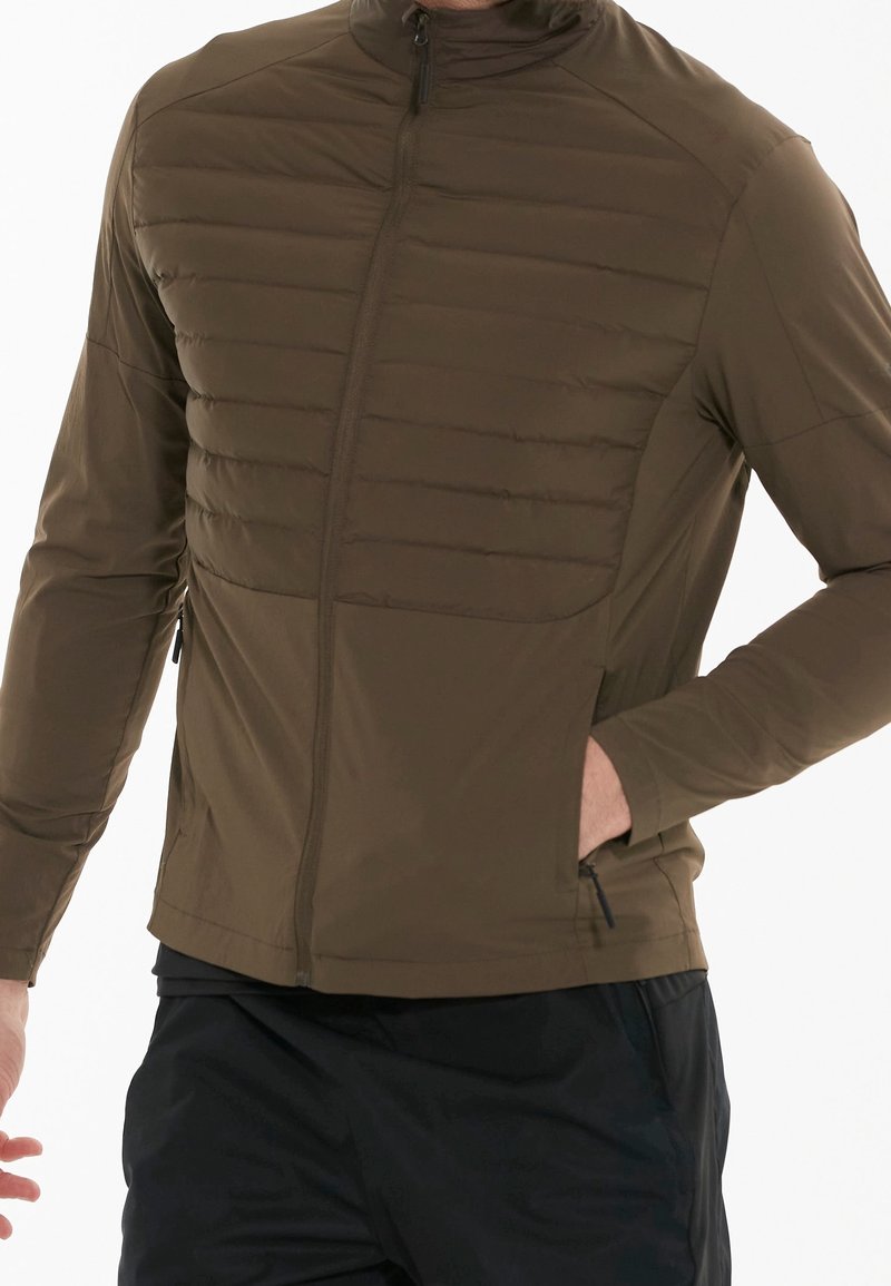 Man wearing an olive green quilted jacket with a front zipper, side pockets, and black pants, hand in jacket pocket.