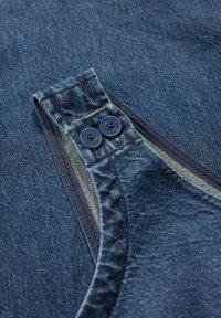 Denim tkanina v temno modri barvi vključuje žep z dvema temno modrima gumboma in kontrastnim šivom ob robovih.