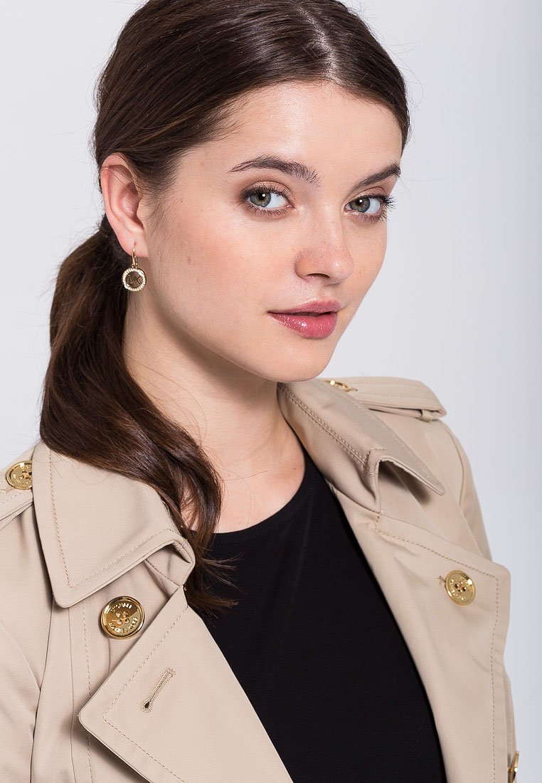 Trench coat beige chiaro con bottoni dorati, indossato sopra un top nero. Il modello ha i capelli di lunghezza media e indossa orecchini dorati a cerchio.