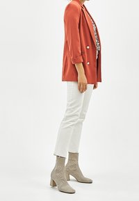 Blazer v rezavé barvě se dvojitými knoflíky, doplněný bílými kalhotami a béžovými ponožkovými botami, předvádějící texturovaný materiál a válcové podpatky.