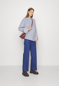 Un pantalon large bleu associé à une chemise à rayures à boutons. Un petit sac à main bordeaux complète la tenue, avec des bottes noires épaisses.