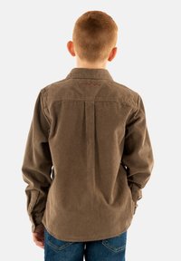 Camisa de pana marrón de manga larga con cuello abotonado, que presenta un pliegue en el yugo trasero y un sutil detalle de costura roja. Llevada con vaqueros azules.