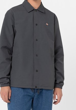 Mężczyzna noszący ciemnoszarą kurtkę zapinaną na guziki z elastycznymi mankietami i dwoma bocznymi kieszeniami, w zestawieniu z niebieskimi dżinsami.