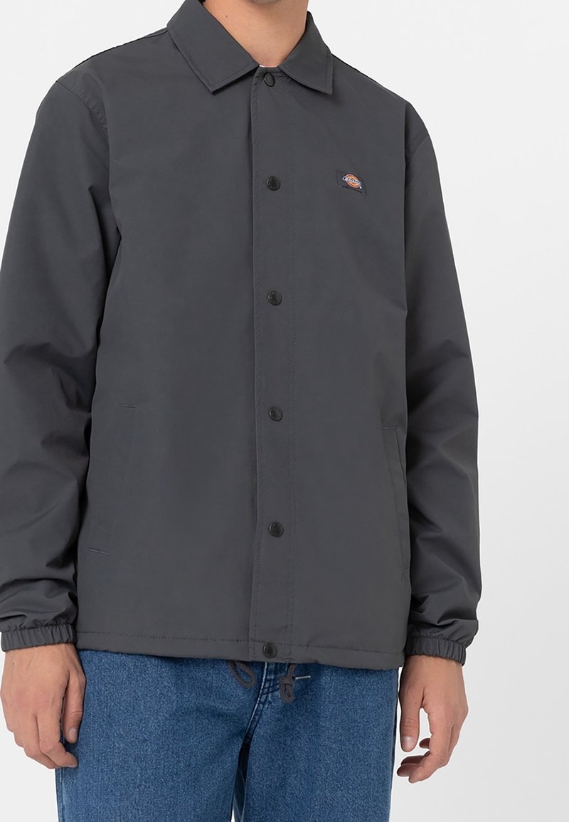 Homme portant une veste boutonnée gris foncé avec des poignets élastiques et deux poches latérales, associée à un jean en denim bleu.