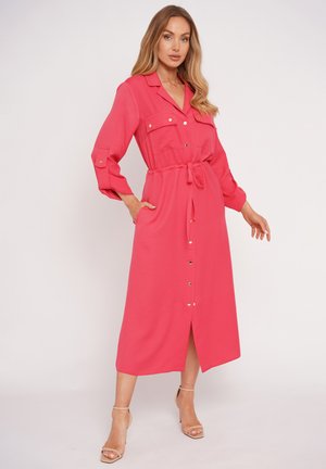 Kobieta prezentująca koralowo-różową sukienkę midi z długim rękawem, zapinaną na guziki z przodu, z kieszeniami na piersi, paskiem w talii oraz beżowymi sandałami na obcasie.