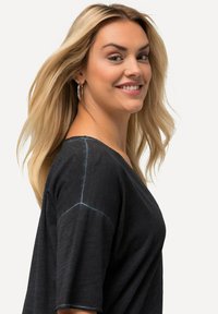 Černý volný top s V-výstřihem, krátkými rukávy a kontrastním modrým stehem podél švů. Nosí ho usmívající se osoba.