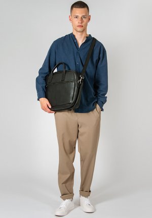 Black leather briefcase with top handles and a detachable shoulder strap. Smooth texture and minimalist design. Model wearing casual attire.