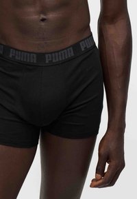 Αρσενικός κορμός που φοράει μαύρα boxer Puma με λογότυπο στη μέση, εμφανίζοντας την κοιλιά και το δεξί χέρι σε απλό φόντο.