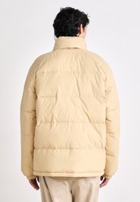 Puffer jacket in light beige, featuring horizontal paneling, high collar, and elastic cuffs, made from smooth, insulated fabric.