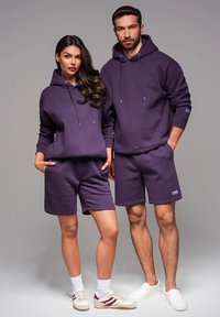 Des sweatshirts à capuche violets et des shorts en tissu lisse, dotés de cordons de serrage et d'accents de logo, portés avec des baskets contrastantes.