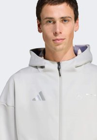 Hellgraue Kapuzenjacke mit glatter Textur, ausgestattet mit einem Reißverschluss, Adidas-Logo und einem gemusterten Innenfutter.