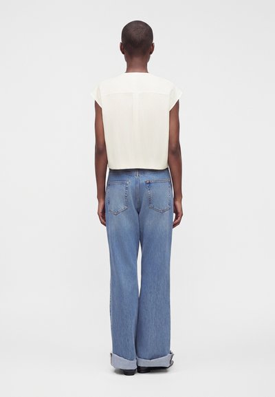 Personne debout, dos à la caméra, portant un haut blanc à manches courtes et un jean bleu large avec revers, sur un fond clair uni.