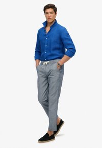 Blauwe linnen shirt met een opstaande kraag, lichtgrijze gestreepte broek en zwarte espadrilles met een jute zool, gedragen met handen in de zakken.
