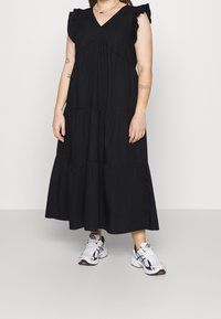 Personne portant une robe maxi noire sans manches à étages avec des épaules à volants et des baskets blanches de sport, debout sur un fond clair uni.