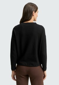 Schwarzer, strukturierter Pullover mit lockerer Passform, langen Ärmeln und weitem Ausschnitt. Von hinten gezeigt, kombiniert mit braunen Hosen.