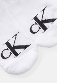 Hvite bomullssokker med en fremtredende svart "ck"-logo. Designet inkluderer en sømløs tå og en myk, teksturert finish.