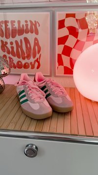 Pelni pelni un baltas sports apavi ar zaļām svītrām uz koka virsmas, blakus disko bumbas lampai un sarkanai retro sienas mākslai.