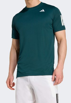 Homme portant un t-shirt de sport à manches courtes vert foncé avec un logo blanc sur la poitrine et la manche, associé à un short blanc avec des rayures noires.