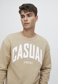 Béžová mikina s bílým nápisem "CASUAL FRIDAY". Má kulatý výstřih a dlouhé rukávy, s měkkou texturou. Jednoduchá, bez vzorů nebo akcentů.