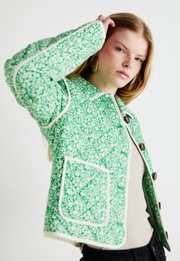 Giacca floreale verde con un motivo testurizzato, contorni bianchi a contrasto, colletto e bottoni grandi; presenta una tasca frontale.