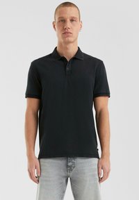 Zwarte polo met een kraag, korte mouwen en een drie-knopen sluiting. Stevige stof, strak ontwerp, gecombineerd met lichtgrijze jeans.