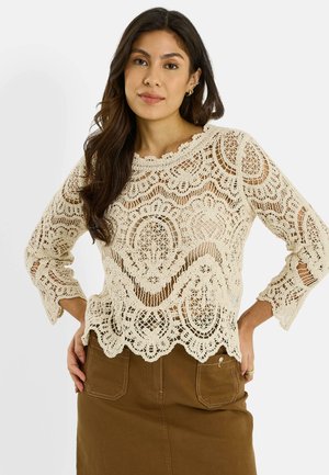 Mujer con cabello largo y oscuro, vistiendo una blusa de encaje de crochet color crema y una falda marrón, con las manos apoyadas en las caderas.