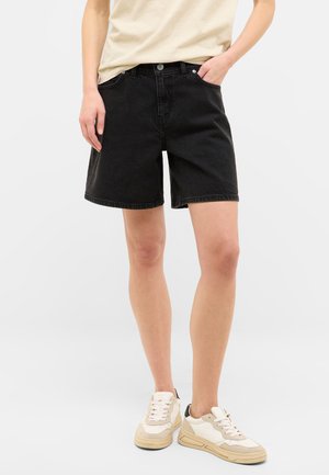Chalecos de mezclilla negra con cinco bolsillos, corte recto y dobladillo cosido. Combinados con zapatillas crema. Diseño y textura minimalistas.