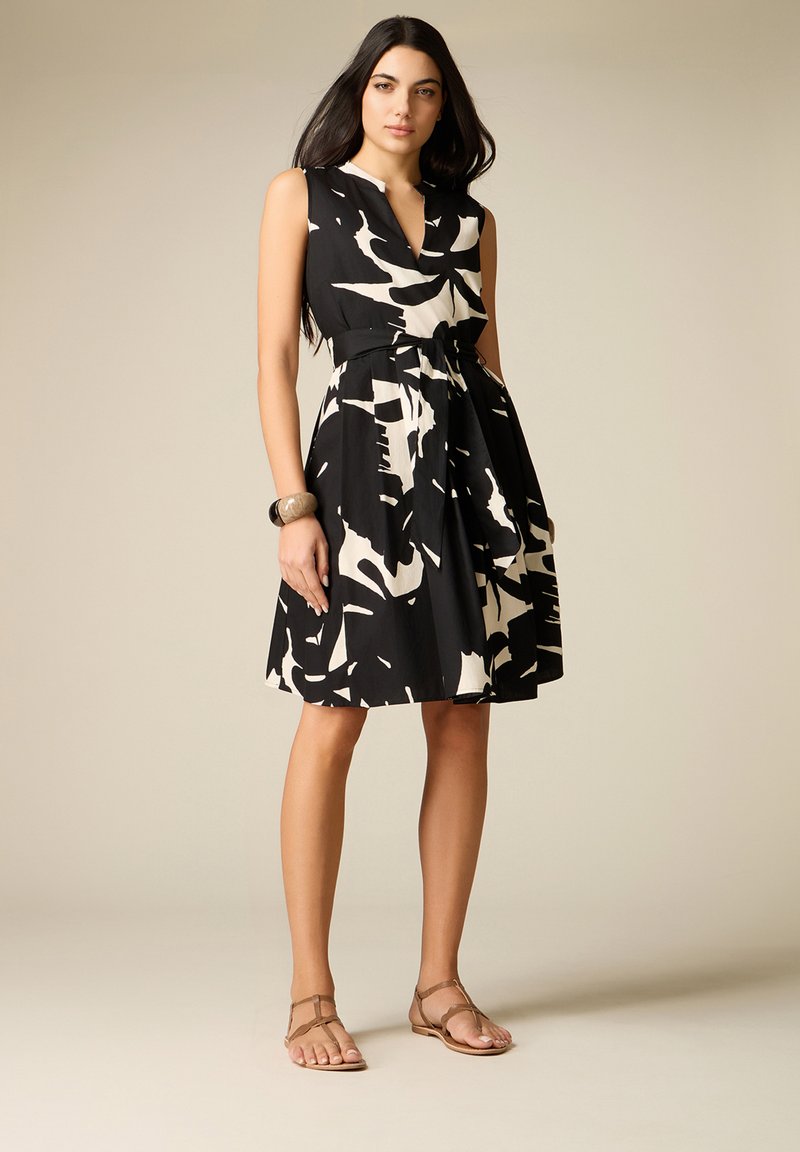 Kobieta w bezrękawowej, czarno-białej sukience o abstrakcyjnym wzorze do kolan, z paskiem, brązowych sandałach i bransoletce, stojąca na jednolitym tle.