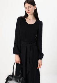 Musta, pitkähihaisuus mekko, jonka bodice-osa on rypytetty ja vyötäröllä on sidottu vyö, yhdistetty mustaan käsilaukkuun.