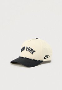 Béžová a černá kšiltovka Nike s vyšitým nápisem "New York" vpředu a logem Nike na boku, umístěná na světle šedém pozadí.