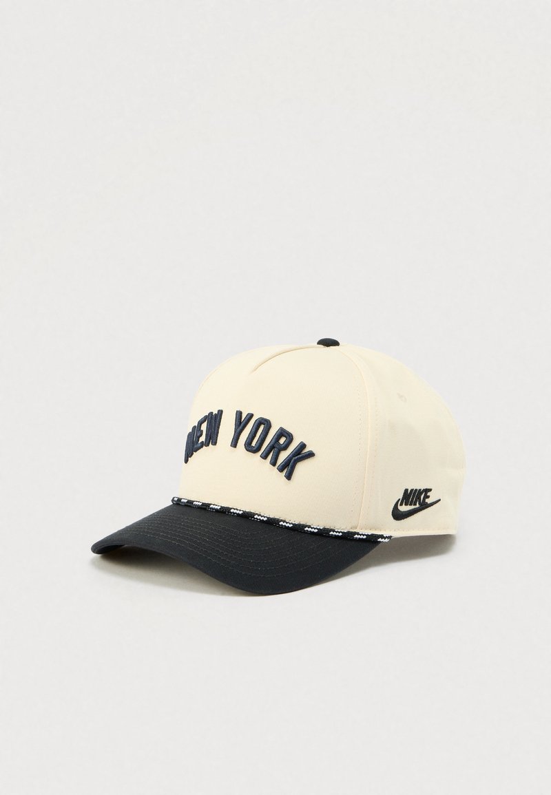 Béžová a černá kšiltovka Nike s vyšitým nápisem "New York" vpředu a logem Nike na boku, umístěná na světle šedém pozadí.