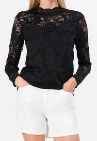 Haut à manches longues en dentelle noire avec motifs floraux, encolure festonnée et finition texturée ; porté avec un short blanc.