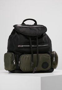 Sac à dos en tissu noir avec cordon de serrage ajustable, plusieurs zips et deux poches latérales vertes. Présente un emblème rond à l'avant.