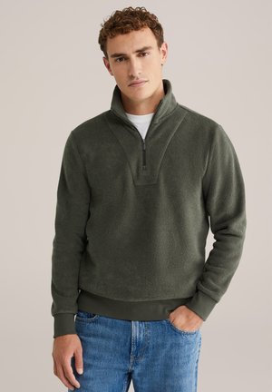 Sudadera de forro polar verde con cuello de media cremallera, superficie texturizada, puños acanalados y color sólido. Combinada con jeans de mezclilla azul.
