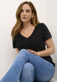 Melna kokvilnas T-kreklu ar V veida kakla izgriezumu un īsām piedurknēm, kopā ar gaiši ziliem džinsiem. Sieviete sēž, atslābinātā pozā, gaišā fona.