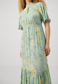 Blumenkleid aus leicht grünem Stoff mit gelben und lila Blumenmustern, kurzen Ärmeln, einem hohen Halsausschnitt und einem gestuften Rockdesign.