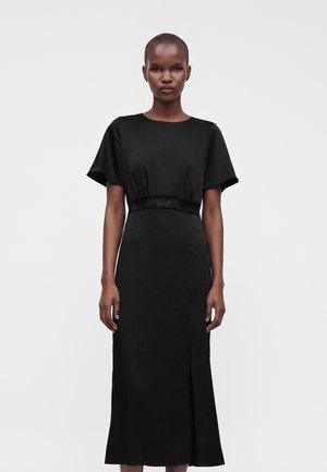 Femme aux cheveux courts porte une robe midi noire à manches courtes avec une fente, debout devant un fond clair uni.