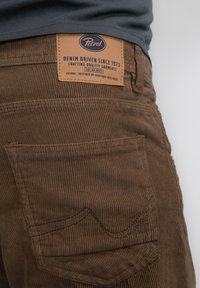 Bordeaux corduroy broek met verticale ribbels, voorzien van een tan leren patch op de taille met merk- en herkomstdetails, en achterzakken.