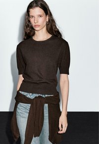 Pull court en tricot marron à manches courtes avec un col rond, associé à un jean bleu clair et une étole marron, nouée à la taille.