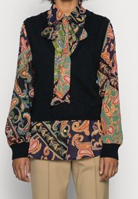 Czarny dzianinowy kamizelka nałożona na bluzkę w wielokolorowy wzór paisley z szerokimi rękawami. W szczegółach znajduje się duża kokarda przy kołnierzyku oraz kontrastowe mankiety.
