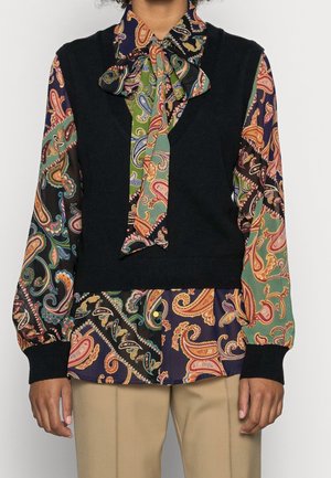 Sort strikket vest over en bluse med flere farver i paisley-mønster og brede ærmer. Detaljer inkluderer en stor sløjfe ved kraven og kontrasterende manchetter.