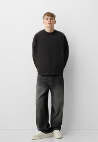 Musta pitkähihaine sweatshirt yhdistettyinä löysän mallisiin haalistuneisiin harmaisiin farkkuihin ja valkoisiin lenkkareihin, taustalla on yksinkertainen valkoinen tausta.