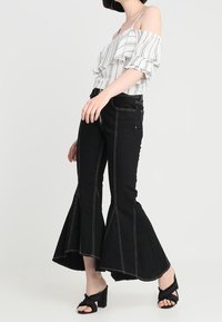 Top bianco a righe con spalle scoperte e dettagli a volant, abbinato a jeans neri a vita alta con zampa d'elefante e cuciture a contrasto, dal taglio pulito.