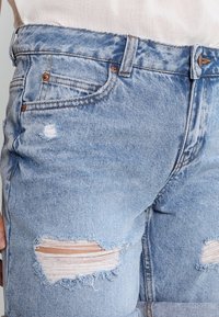 Ljusblå jeans i denim med slitna detaljer, inklusive revor på knäna, kopparfärgade knappdetaljer och klassisk design med fem fickor.