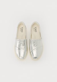 Silberne metallische Slip-On-Schuhe mit einem Kreuzdesign, ausgestattet mit einem weichen, strukturierten Obermaterial und einer cremefarbenen Gummisohle. "TOMS"-Branding im Inneren.