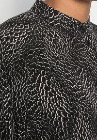 Musta paita korkealla kauluksella, jossa on valkoinen aaltoileva kuvio. Siinä on mustat nappikoristeet ja sileä kangastuntuma.