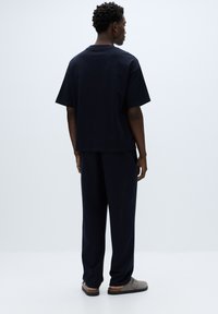 Tmavomodré nadrozmerné tričko s krátkymi rukávmi, v kombinácii s rovnako farebnými voľnými nohavicami a sivými slip-on topánkami, zobrazené zozadu.